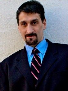 Albert Rizzi - My Blind Spot - Headshot Photo. Albert has dark hair and a dark goatee. He is wearing a navy suit with a blue shirt and a striped necktie